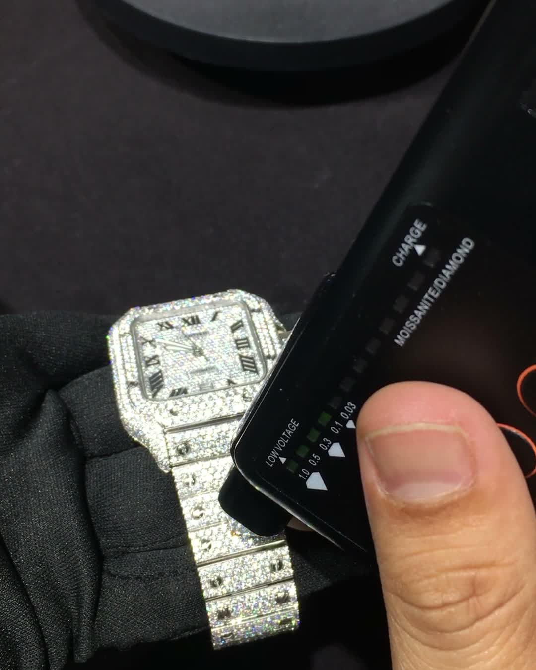 A bench tester probe pressed against the iced case of a fully set watch, with the tester's readout label showing moissanite/diamond and a green indicator lit